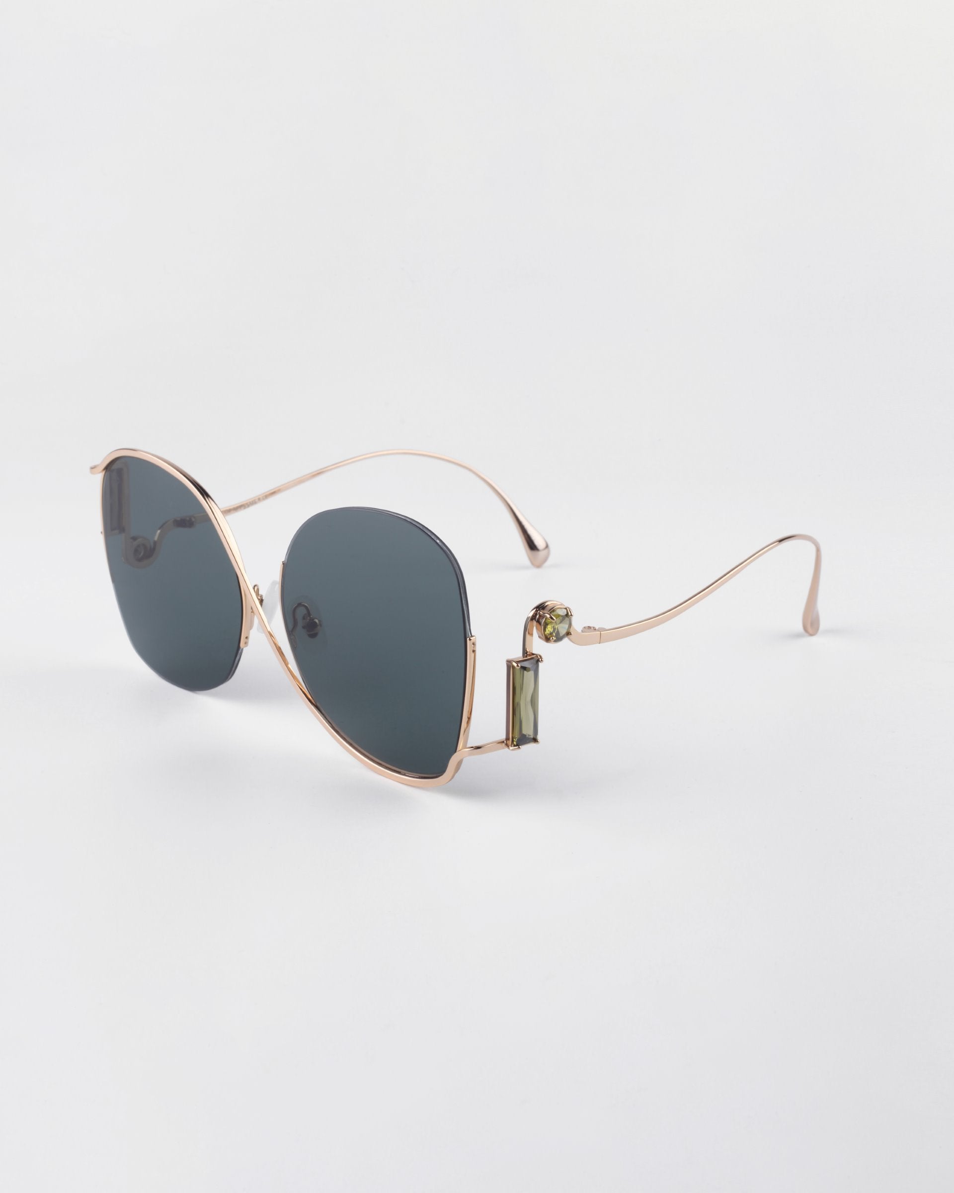 A pair of luxurious sunglasses featuring gold metal frames and dark, UV-protected lenses. The design includes round lenses and thin, curved temple arms with small green decorative details near the hinges. Crystal embellishments add a touch of elegance against a plain white background. These are the Sapphire sunglasses by For Art&#39;s Sake®.