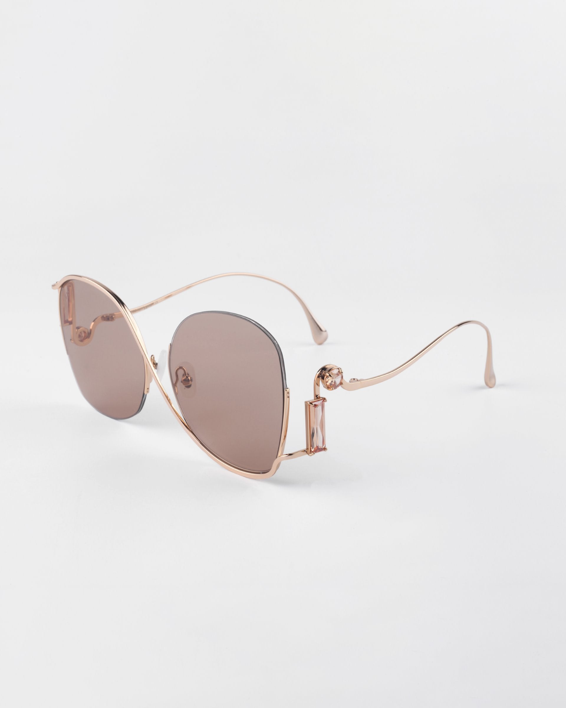 A pair of luxurious For Art&#39;s Sake® Sapphire sunglasses with rose gold frames and brown-tinted lenses. The design features smooth, curved lines with thin temples, and small crystal embellishments on the sides. These stylish glasses with UV protection rest on a white surface.