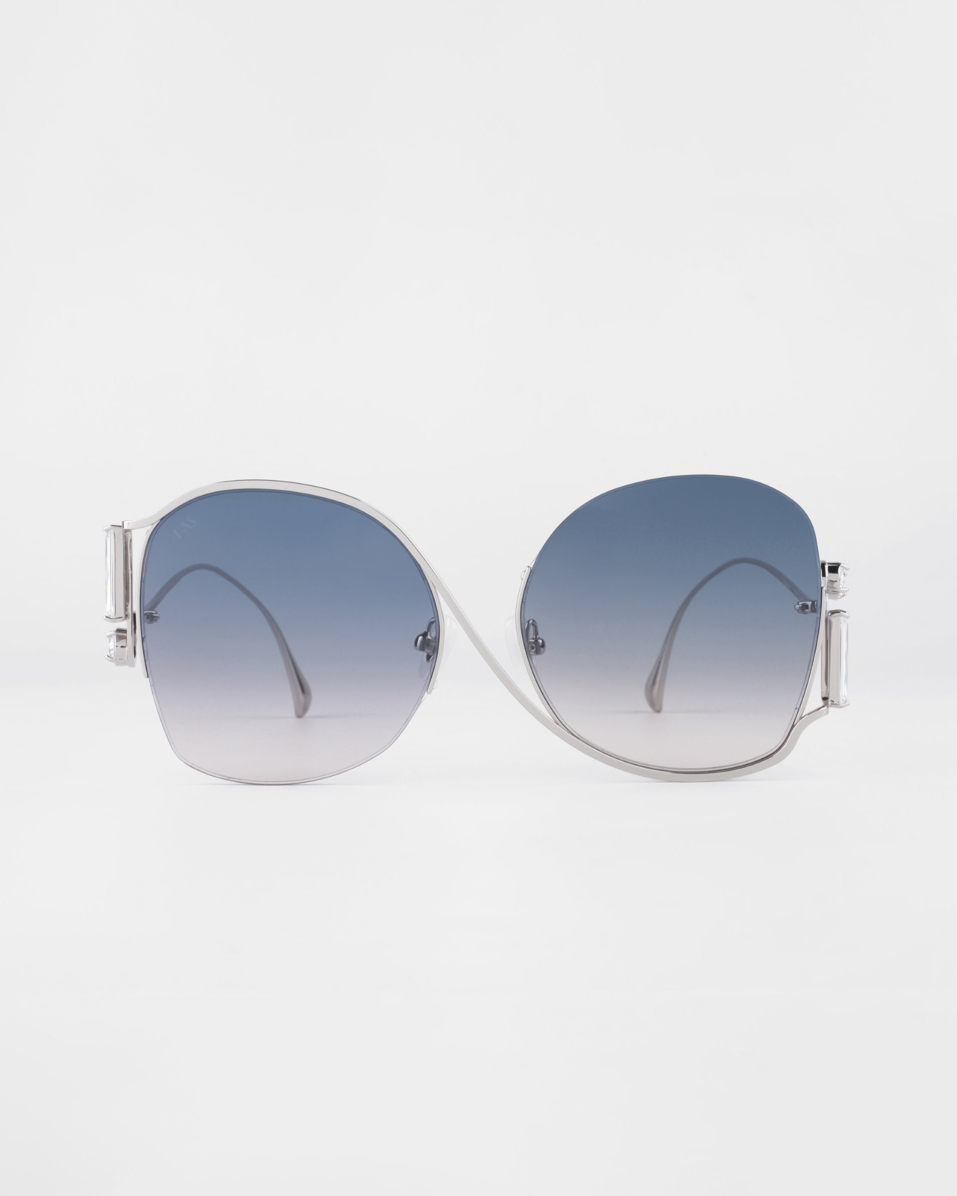 A pair of luxurious For Art&#39;s Sake® Sapphire sunglasses with silver frames and large, gradient lenses. The lenses transition from dark blue at the top to lighter shades towards the bottom. Crystal embellishments add a touch of elegance. The background is white, making the sunglasses the focal point.