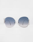 A pair of luxurious For Art's Sake® Sapphire sunglasses with silver frames and large, gradient lenses. The lenses transition from dark blue at the top to lighter shades towards the bottom. Crystal embellishments add a touch of elegance. The background is white, making the sunglasses the focal point.