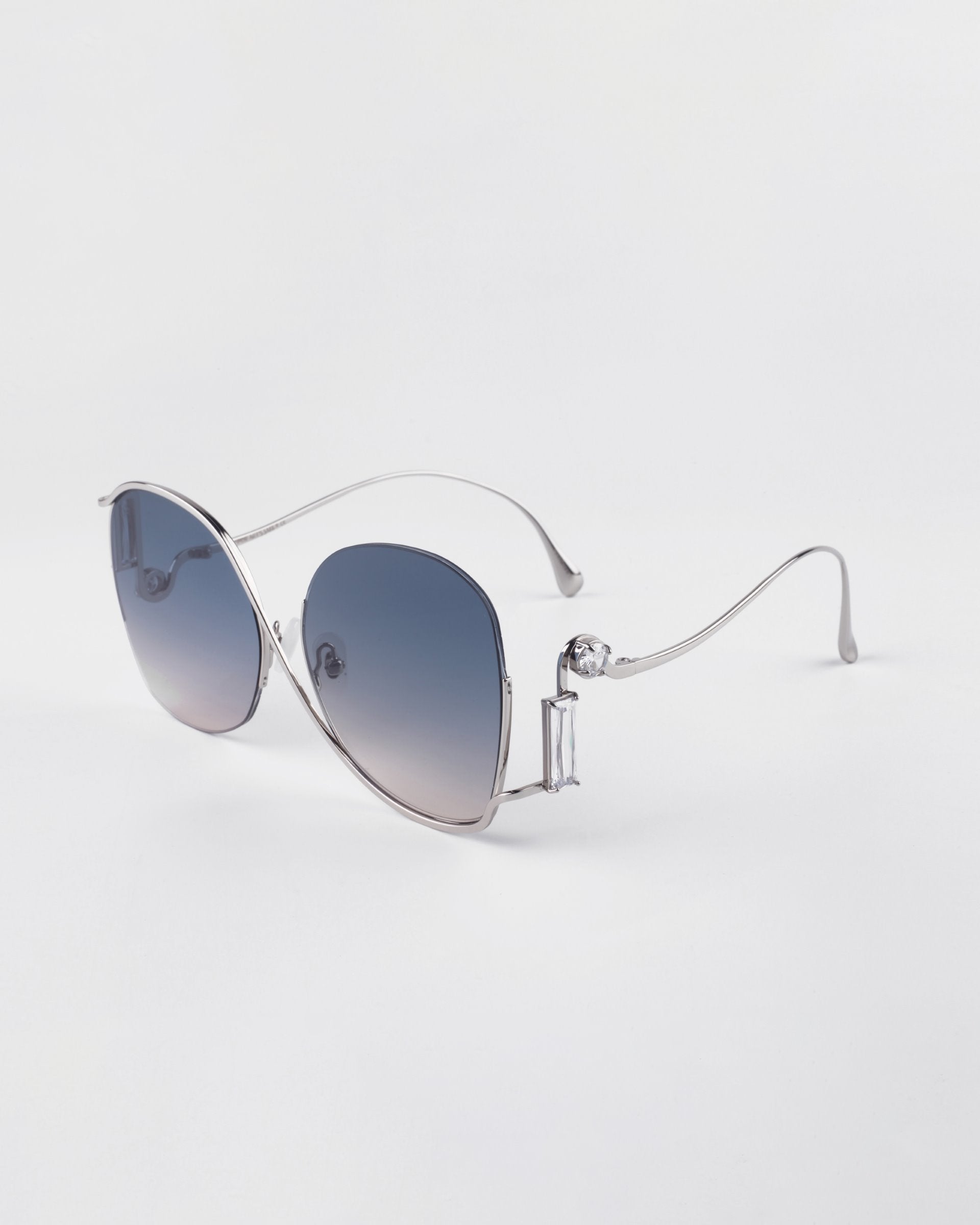 A pair of luxurious silver-framed Sapphire sunglasses from For Art&#39;s Sake® with dark gradient lenses is displayed against a plain white background. The sunglasses have a sleek, modern design with thin temples and slightly oversized lenses, offering exceptional UV protection for your eyes.