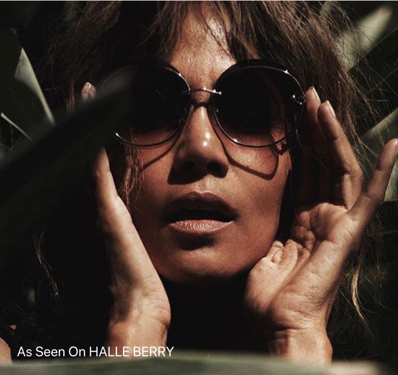 A person wearing dark round Quartz sunglasses with nylon lenses from For Art&#39;s Sake® and holding their hands to their face is partly shadowed by surrounding foliage. Text on the image reads, &quot;As Seen On HALLE BERRY.