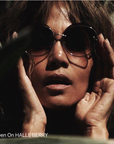 A person wearing dark round Quartz sunglasses with nylon lenses from For Art's Sake® and holding their hands to their face is partly shadowed by surrounding foliage. Text on the image reads, "As Seen On HALLE BERRY.