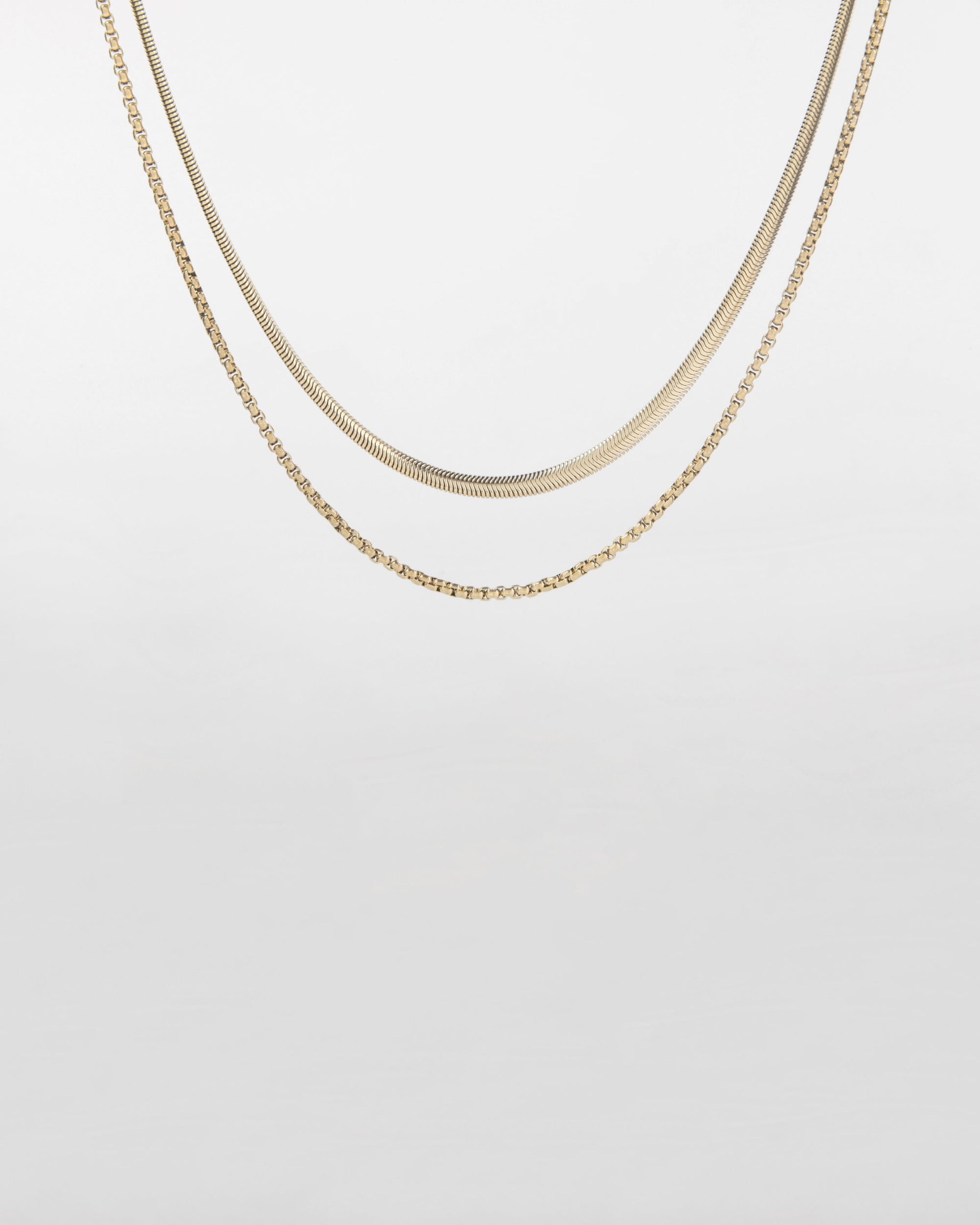 Two delicate gold chains are depicted on a white background. The upper chain is For Art's Sake®'s Snake Glasses Chain with a smooth, flat design, while the lower chain features a more intricate, rope-like pattern. Both chains hang in a gentle curve.