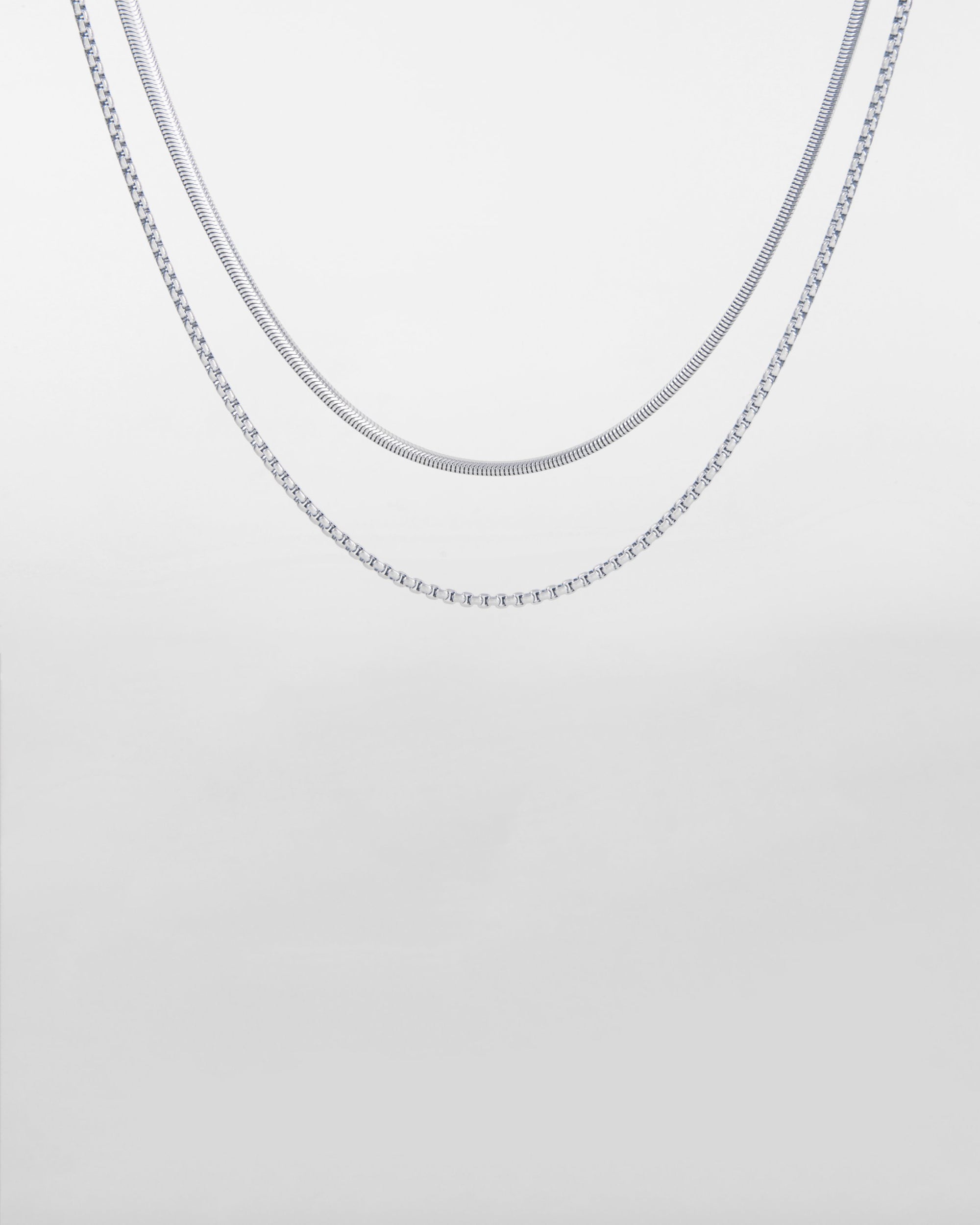 Two delicate, silver Snake Glasses Chains by For Art's Sake® are displayed against a plain, light grey background. The chains have different styles: one is a simple, sleek design while the other features a slightly more textured pattern reminiscent of a gold-plated eyewear chain. Both are draped in a gentle curve.