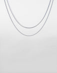 Two delicate, silver Snake Glasses Chains by For Art's Sake® are displayed against a plain, light grey background. The chains have different styles: one is a simple, sleek design while the other features a slightly more textured pattern reminiscent of a gold-plated eyewear chain. Both are draped in a gentle curve.