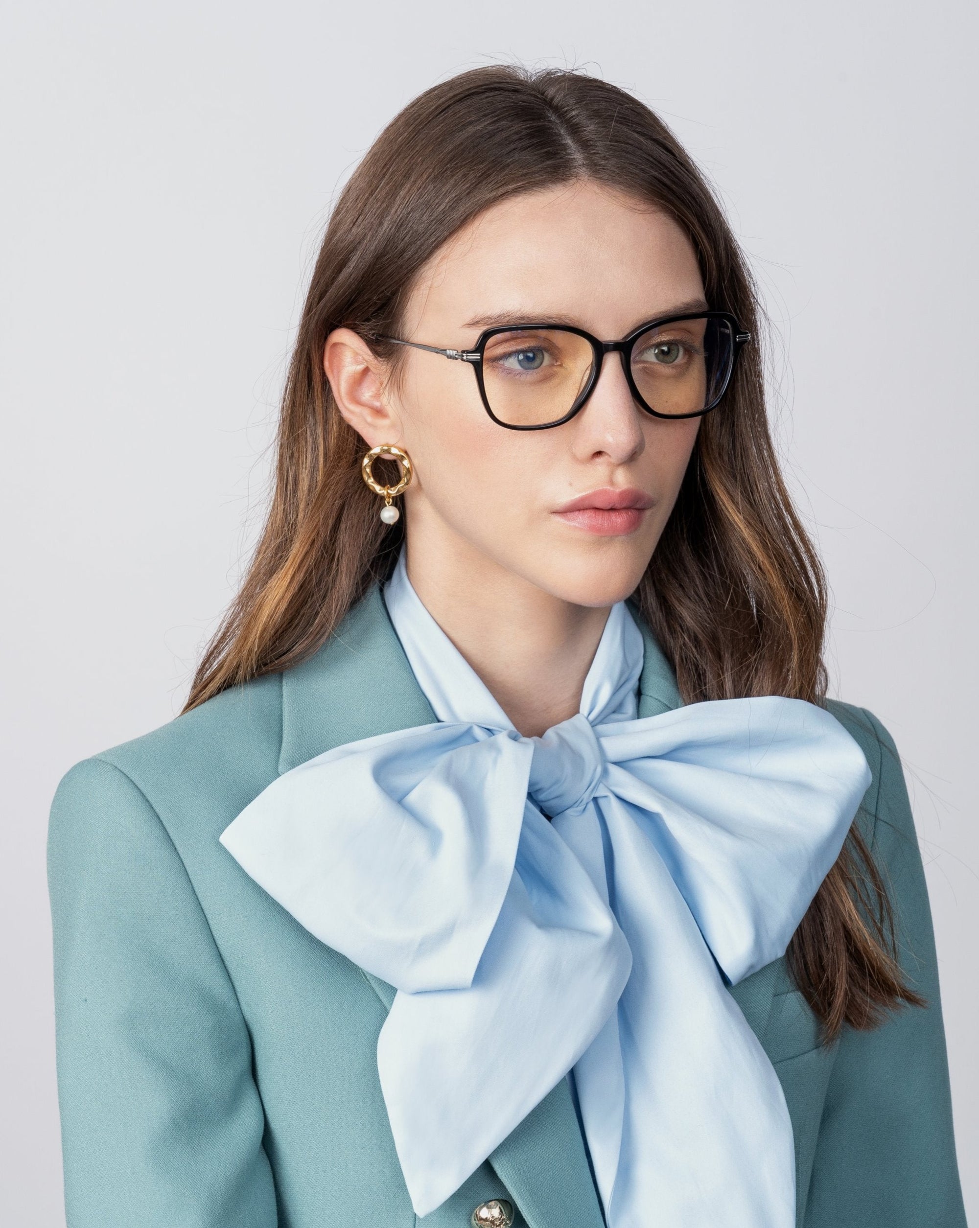A person with long brown hair is wearing For Art's Sake® Sonnet black framed glasses with blue light filters, gold hoop earrings, and a light blue blouse with a large matching bow at the neck. They are also dressed in a teal blazer with a serious expression, looking slightly to the side.
