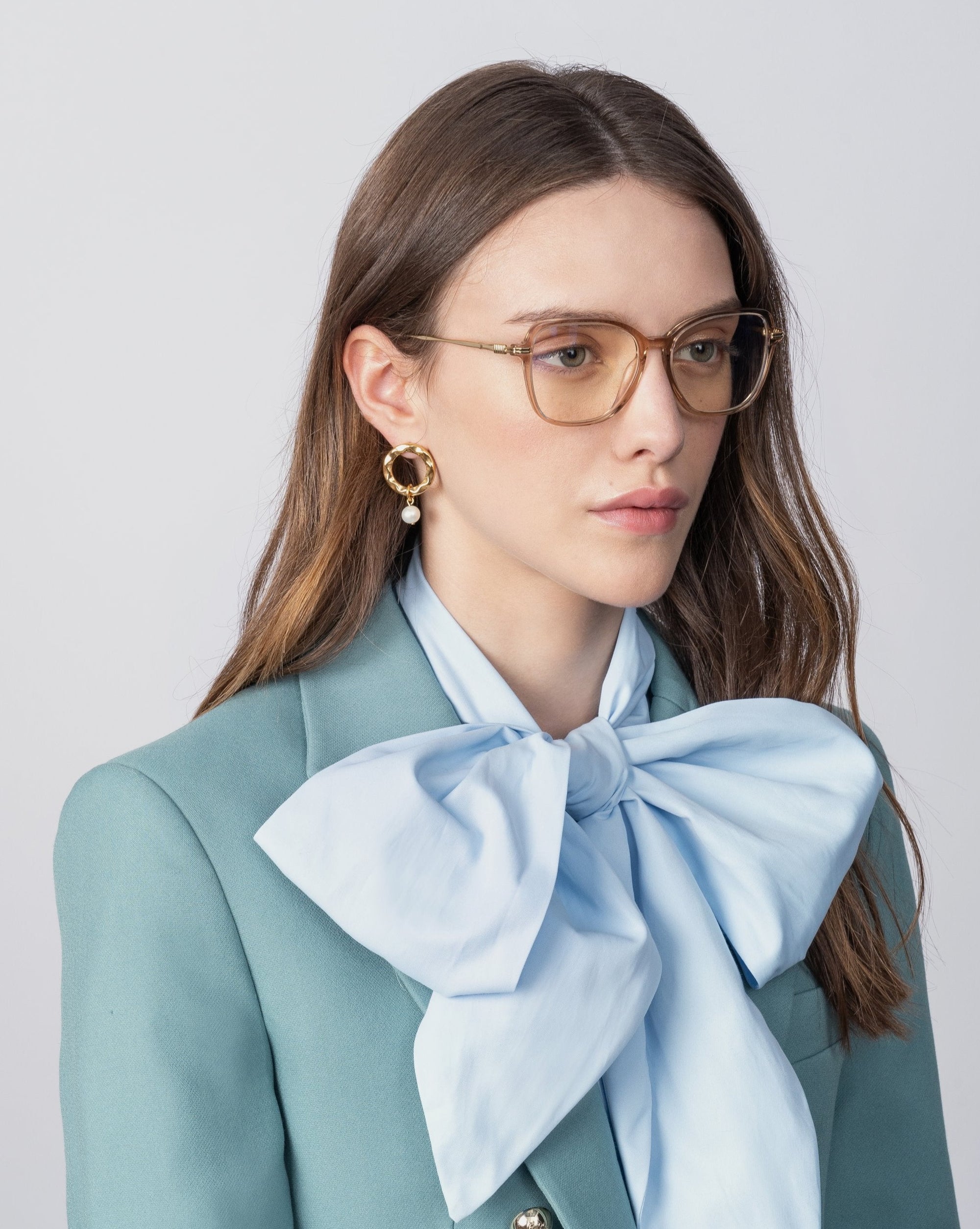 A woman with long brown hair wears transparent Sonnet glasses with blue light filter lenses by For Art&#39;s Sake®, a light teal blazer, and a large light blue bow blouse. She also has large, 18-karat gold-plated earrings. The background is plain and light-colored.
