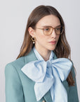 A woman with long brown hair wears transparent Sonnet glasses with blue light filter lenses by For Art's Sake®, a light teal blazer, and a large light blue bow blouse. She also has large, 18-karat gold-plated earrings. The background is plain and light-colored.