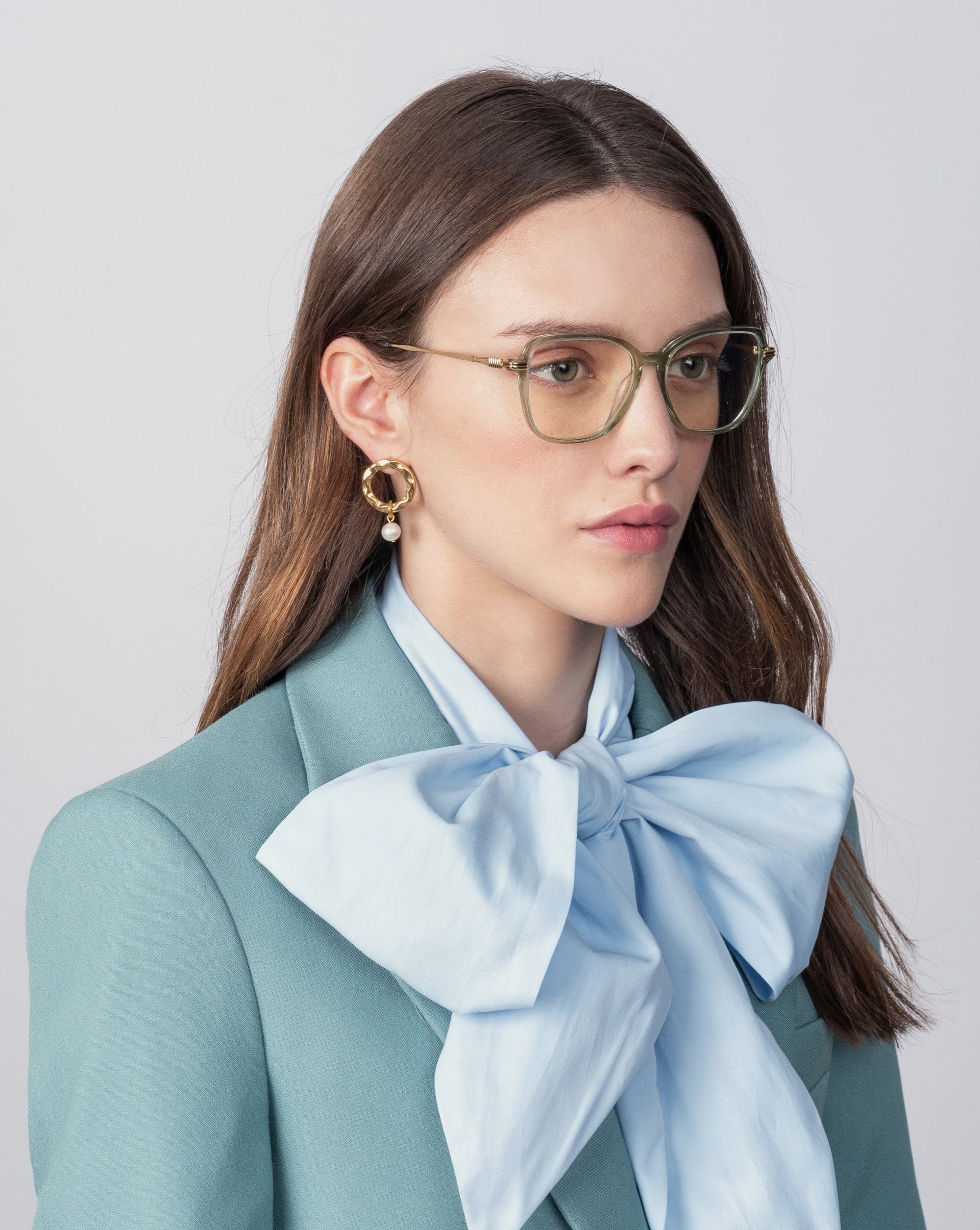 A woman with long brown hair, wearing Sonnet by For Art&#39;s Sake® and a pair of 18-karat gold-plated earrings, is pictured against a plain backdrop. Dressed in a blue blouse with an oversized bow at the neck and a teal blazer, her expression remains neutral.
