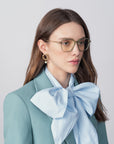 A woman with long brown hair, wearing Sonnet by For Art's Sake® and a pair of 18-karat gold-plated earrings, is pictured against a plain backdrop. Dressed in a blue blouse with an oversized bow at the neck and a teal blazer, her expression remains neutral.