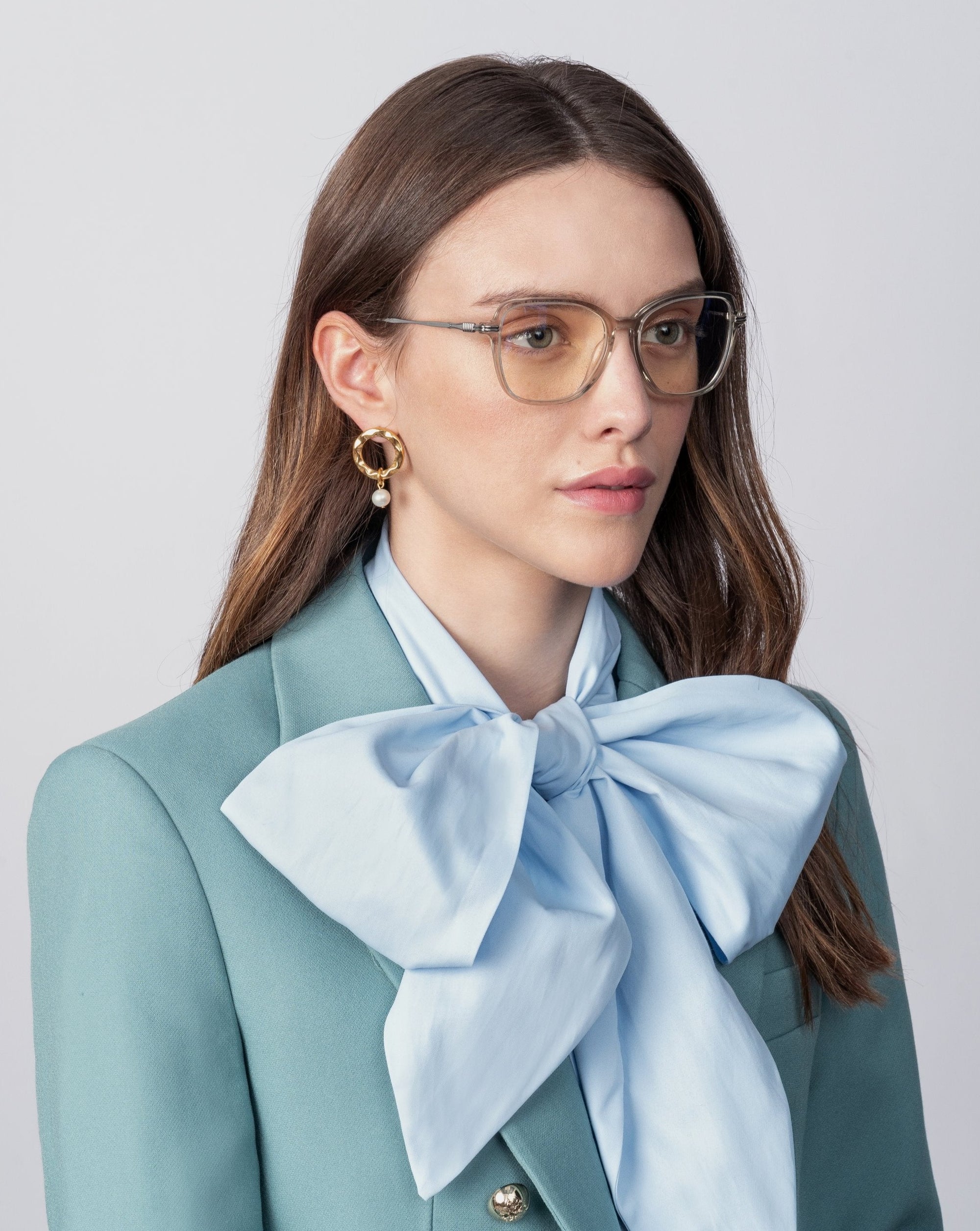 A person with long brown hair and glasses with prescription lenses is shown from the shoulders up, wearing a green blazer and a large light blue bow tie. They have 18-karat gold-plated earrings by For Art&#39;s Sake® Sonnet and a neutral expression against a plain light background.