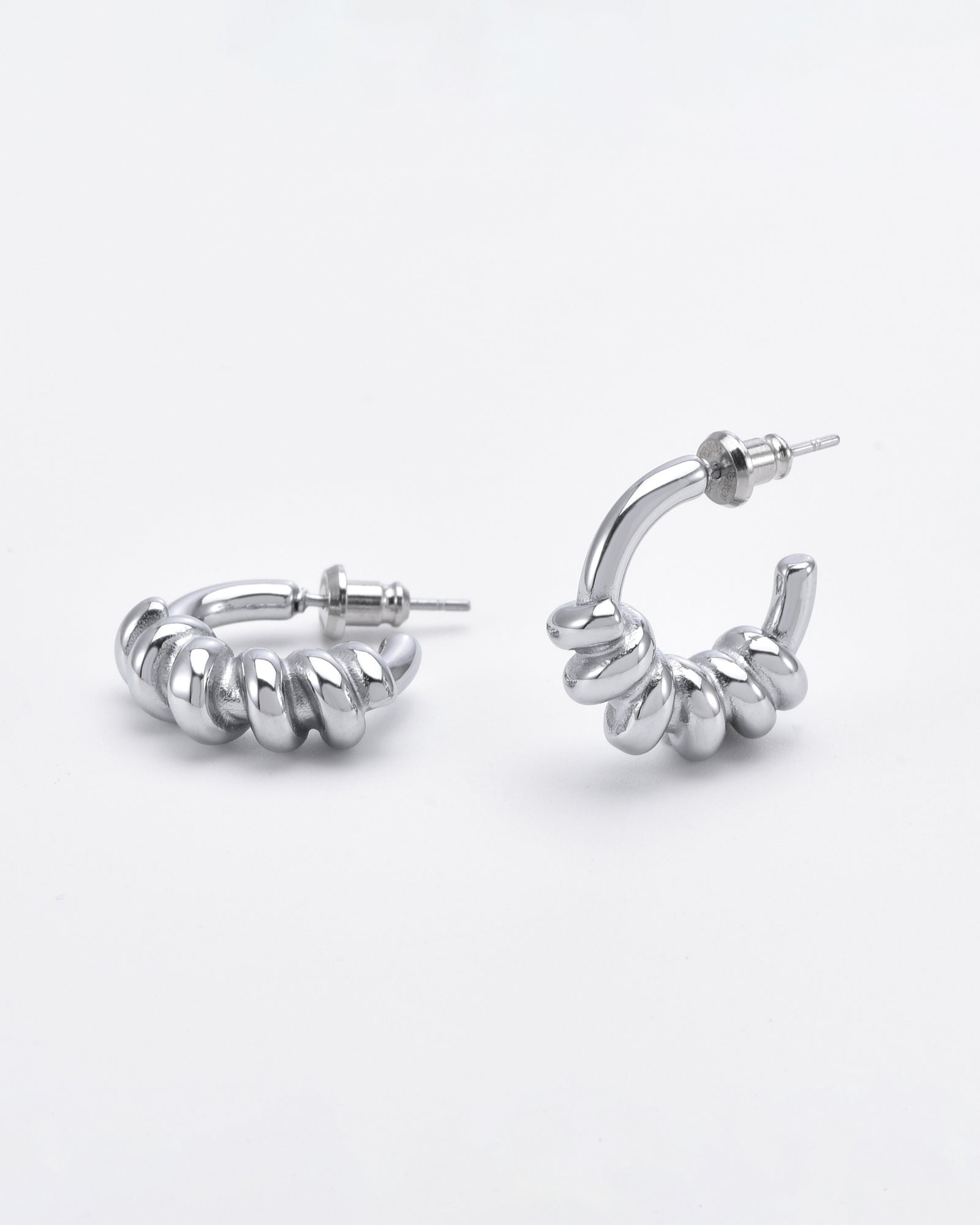 A pair of Swirl Earrings Silver from For Art's Sake® are displayed on a white background. One earring is standing upright, while the other lies on its side, showcasing their shiny and polished finish.
