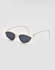 The For Art's Sake® Ornate sunglasses feature gold-tone frames and dark, oval-shaped lenses. The design includes thin temples and intricate detailing near the hinges, creating a sleek and elegant appearance. Plated in 18-karat gold, they offer 100% UVA & UVB protection. The sunglasses are set against a plain background to emphasize their sophisticated design.