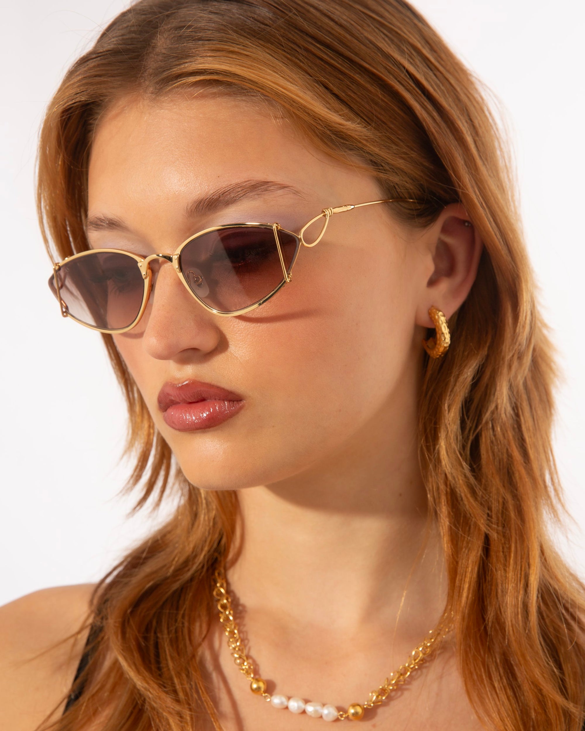 A person with shoulder-length light brown hair wears stylish Ornate sunglasses by For Art&#39;s Sake®, a gold hoop earring, and an 18-karat gold-plated necklace accented with pearls. The background is plain white, highlighting the accessories and the person&#39;s relaxed expression.