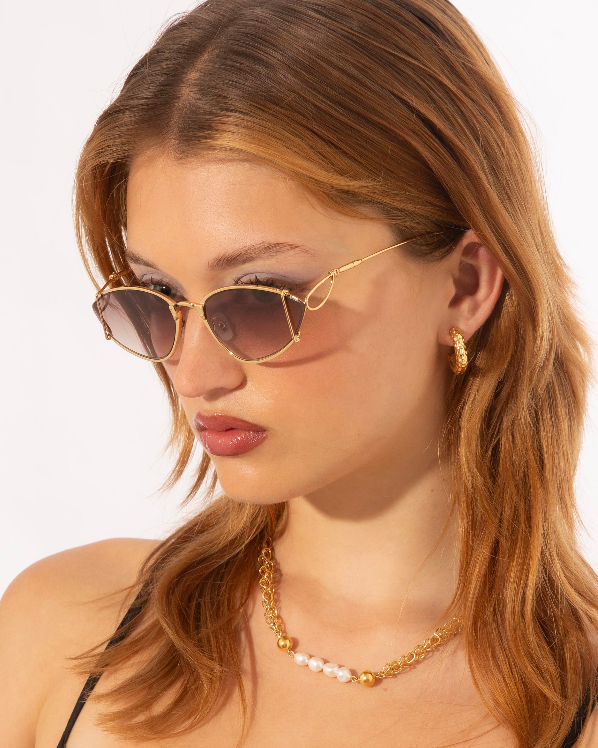 A person with long light brown hair is wearing 18-karat gold-plated, geometric For Art's Sake® Ornate sunglasses and gold jewelry, including a necklace with a mix of gold and white beads and gold hoop earrings. The individual is looking slightly down and to the side against a white background.