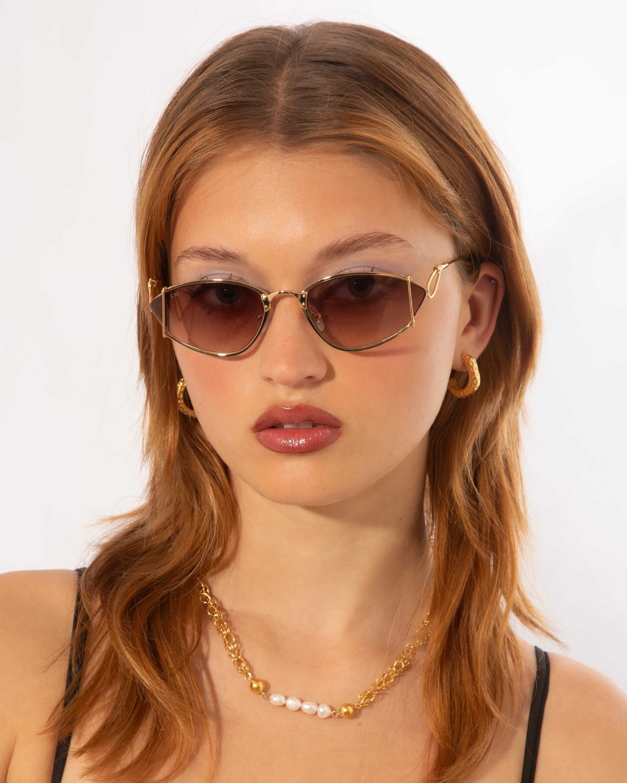 A person with medium-length brown hair wears fashionable almond-shaped For Art's Sake® Ornate sunglasses with tinted lenses, gold hoop earrings, and an 18-karat gold-plated stainless steel chain necklace with pearl accents. The background is a plain white setting.