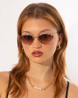 A person with medium-length brown hair wears fashionable almond-shaped For Art's Sake® Ornate sunglasses with tinted lenses, gold hoop earrings, and an 18-karat gold-plated stainless steel chain necklace with pearl accents. The background is a plain white setting.