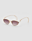 A pair of Ornate sunglasses by For Art's Sake® featuring stylish, vintage-style gold-tone metal frames and tinted lenses. The arms are slightly curved, and the lens shape is subtly cat-eye. Offering 100% UVA & UVB protection, the sophisticated design delivers a modern yet elegant look against a plain gray background.