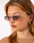 A person with long, light brown hair wearing stylish, angular For Art's Sake® Ornate sunglasses that offer 100% UVA & UVB protection and gold hoop earrings. They have a neutral facial expression and are dressed in a sheer beige top, set against a simple, light background.