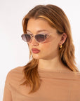 A woman with light brown hair wears For Art's Sake® Ornate geometric sunglasses with rose-tinted, ultra-lightweight nylon lenses providing 100% UVA & UVB protection. She accessorizes with small 18-karat gold-plated pendant earrings and a beige top, looking slightly to her left against a plain white background.