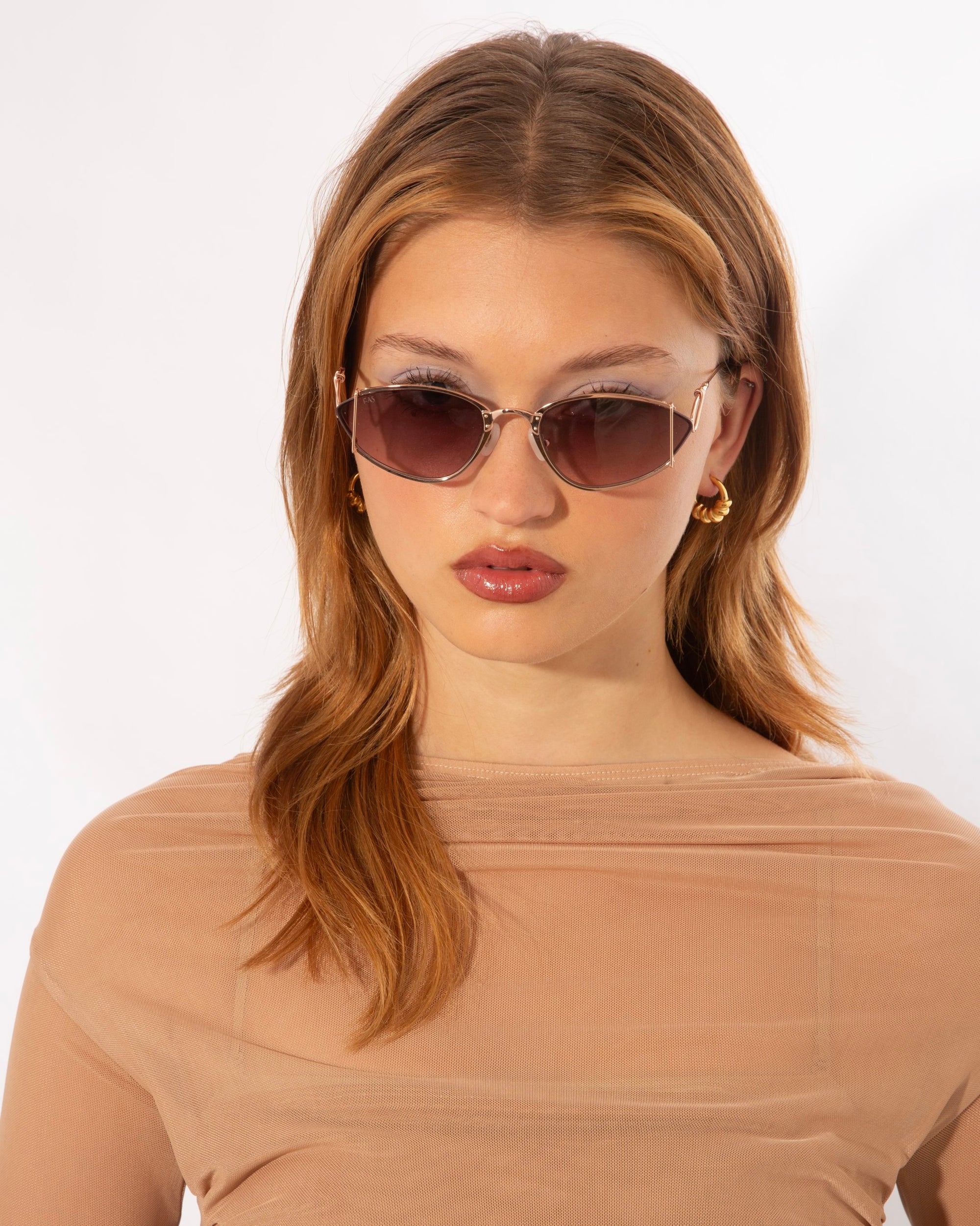 A woman with light brown hair and wearing stylish For Art&#39;s Sake® Ornate sunglasses that offer 100% UVA &amp; UVB protection looks at the camera. She is dressed in a light beige, semi-sheer top, and has gold hoop earrings. The background is plain white.