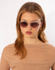 A woman with light brown hair and wearing stylish For Art's Sake® Ornate sunglasses that offer 100% UVA & UVB protection looks at the camera. She is dressed in a light beige, semi-sheer top, and has gold hoop earrings. The background is plain white.