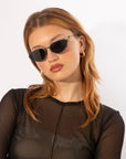 A woman with medium-length, reddish-brown hair is wearing black For Art's Sake® Ornate sunglasses with ultra-lightweight nylon lenses and gold hoop earrings. She has a neutral expression and is dressed in a black, semi-transparent top. The For Art's Sake® Ornate sunglasses offer 100% UVA & UVB protection, and the background is white.