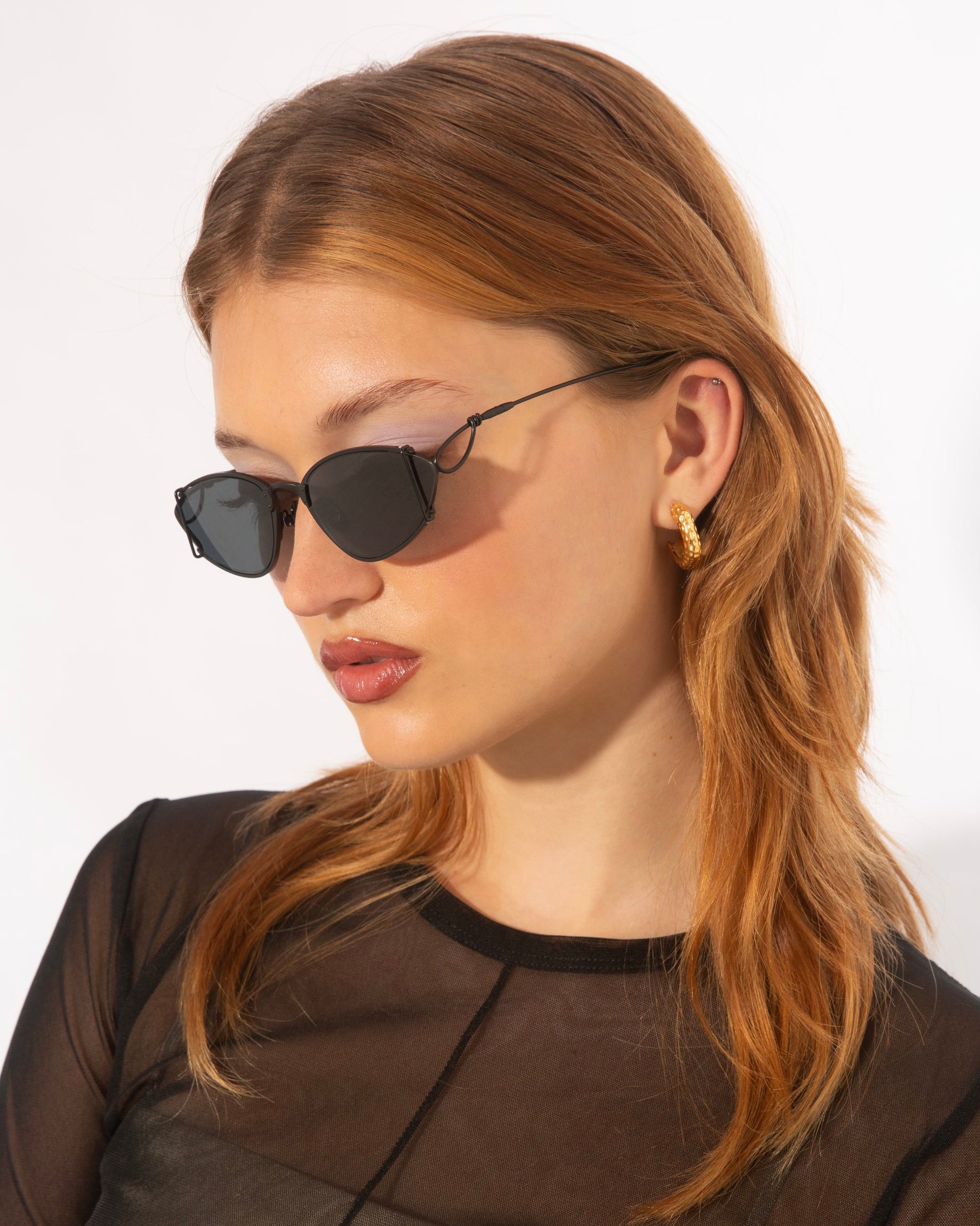 A person with light skin, long auburn hair, and wearing For Art&#39;s Sake® Ornate sunglasses with ultra-lightweight nylon lenses is shown in profile against a plain background. They are dressed in a sheer black top and gold hoop earrings that shimmer like 18-karat gold-plated jewelry.