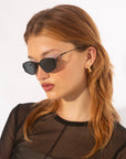 A person with light skin, long auburn hair, and wearing For Art's Sake® Ornate sunglasses with ultra-lightweight nylon lenses is shown in profile against a plain background. They are dressed in a sheer black top and gold hoop earrings that shimmer like 18-karat gold-plated jewelry.