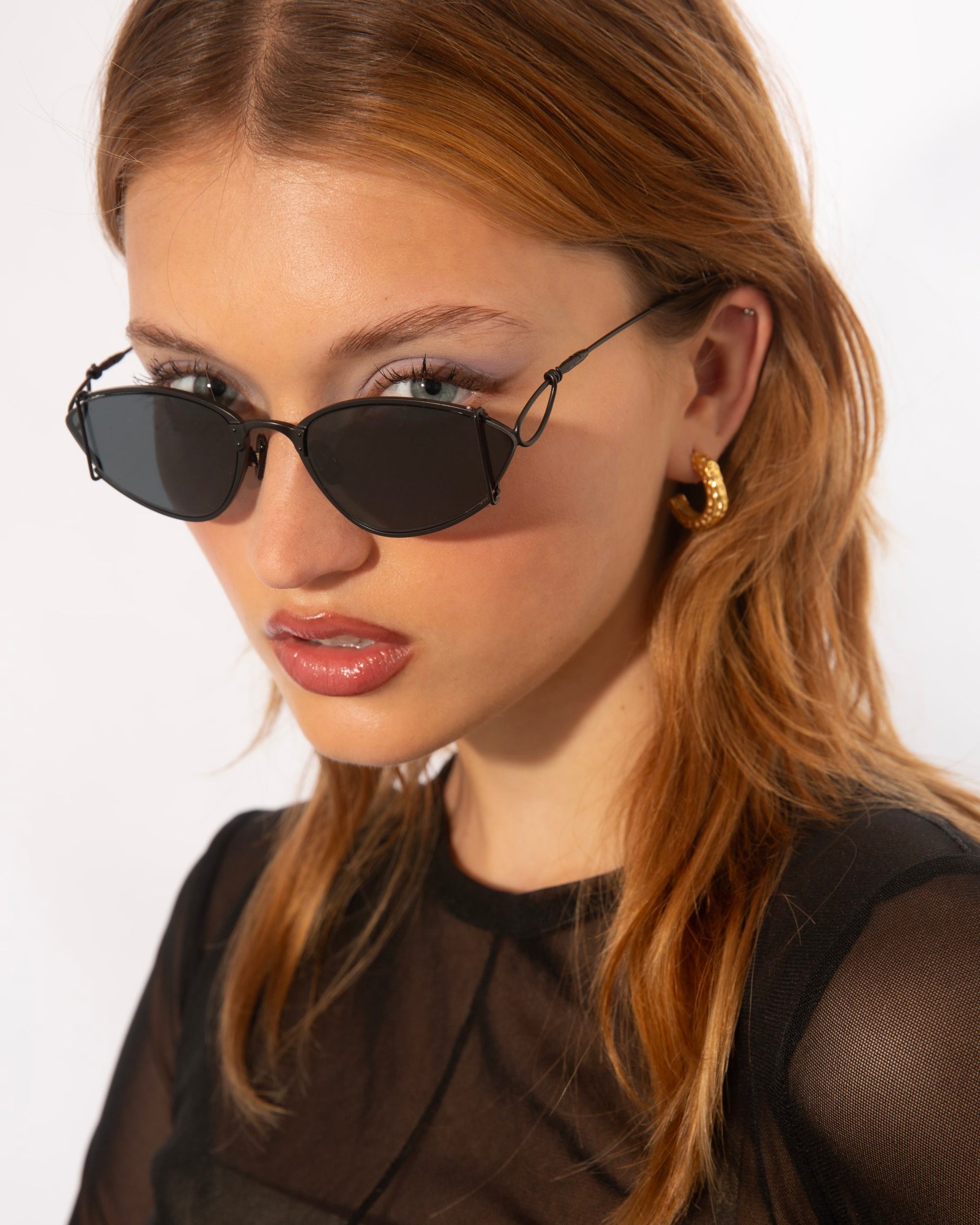 A woman with straight, shoulder-length auburn hair is wearing dark, angular For Art&#39;s Sake® Ornate sunglasses with ultra-lightweight nylon lenses offering 100% UVA &amp; UVB protection and 18-karat gold-plated hoop earrings. She has a serious expression and is dressed in a sheer black top against a plain white background.
