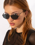 A woman with straight, shoulder-length auburn hair is wearing dark, angular For Art's Sake® Ornate sunglasses with ultra-lightweight nylon lenses offering 100% UVA & UVB protection and 18-karat gold-plated hoop earrings. She has a serious expression and is dressed in a sheer black top against a plain white background.