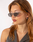 A woman with medium-length light brown hair is wearing dark rectangular sunglasses, specifically the Ornate by For Art's Sake®, with ultra-lightweight Nylon lenses and small round silver earrings. She is looking to the side and has a slight pout on her lips. Her sleeveless top is grey. The background is plain white.