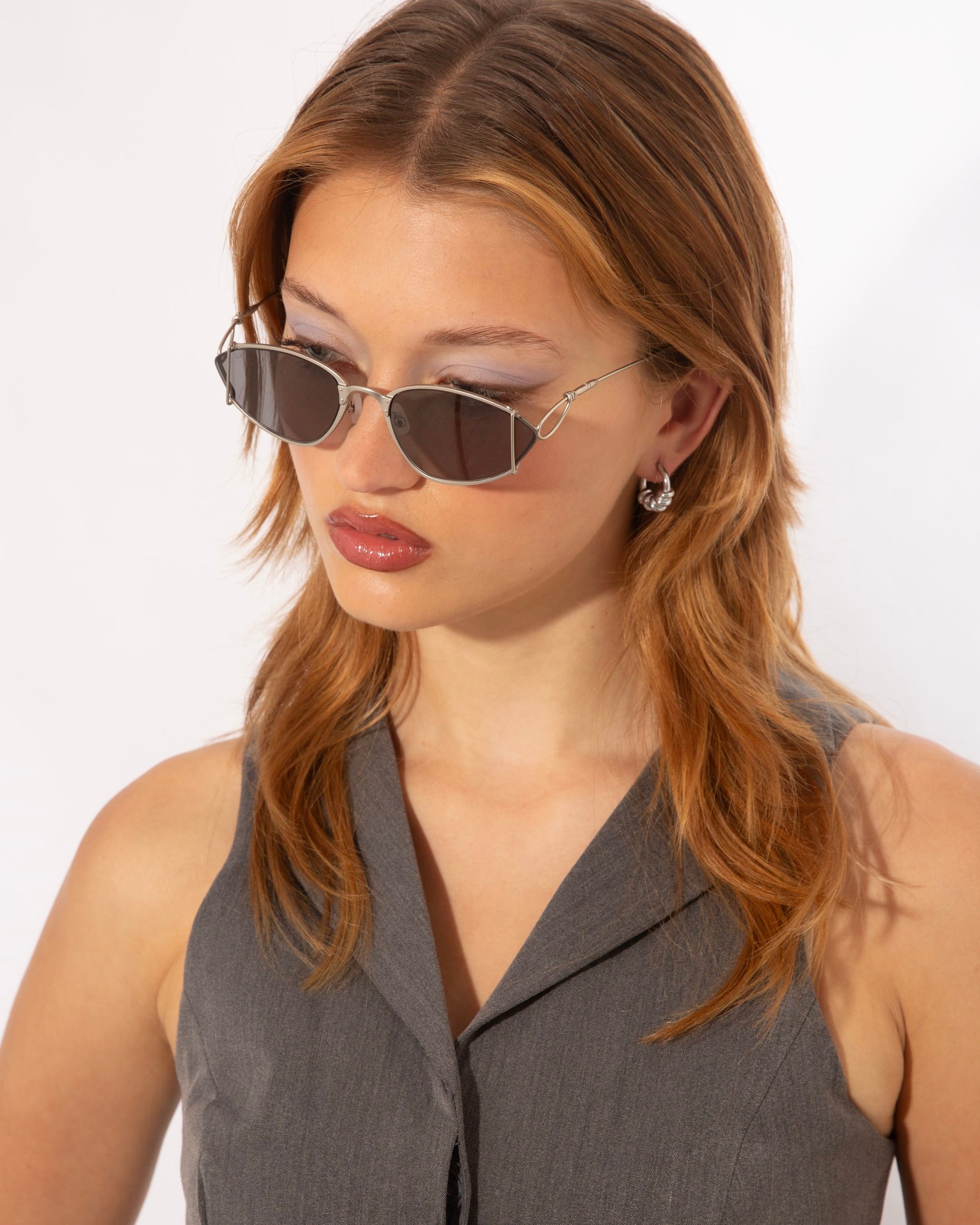 A woman with light brown hair wearing angular For Art&#39;s Sake® Ornate sunglasses featuring ultra-lightweight nylon lenses and 100% UVA &amp; UVB protection looks downward. She has subtle makeup, small hoop earrings, and a sleeveless gray top. The background is plain white.