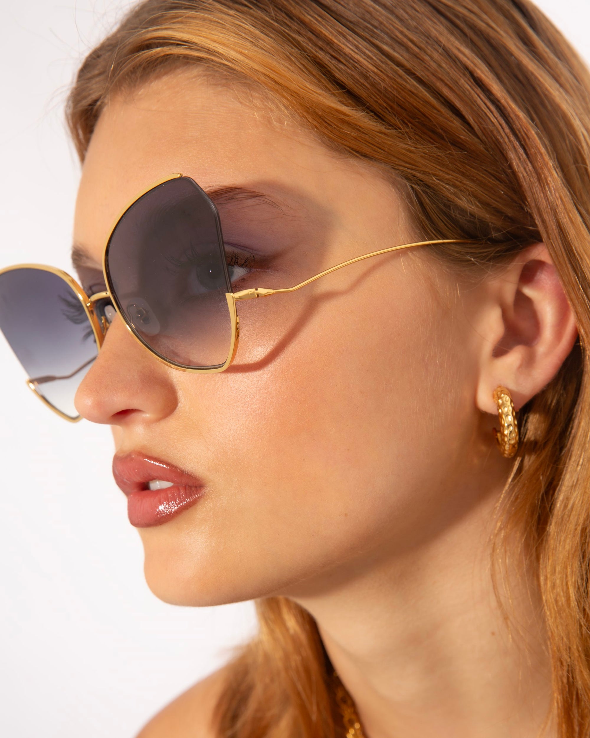 A woman with long, light brown hair is wearing large, dark Watercolour sunglasses by For Art's Sake® and gold-plated stainless steel hoop earrings. Her lips are glossed, and she is gazing to the side. The background is white, making her accessories stand out.