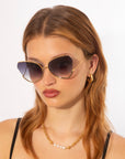 A person with long, light brown hair, wearing stylish Watercolour sunglasses with UV protection by For Art's Sake®, a gold chain necklace with a pearl centerpiece, and gold hoop earrings poses against a plain white background. The individual is dressed in black straps.