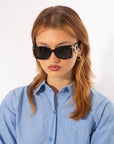 A person with shoulder-length, light brown hair is wearing Sculpture - Black rectangular acetate sunglasses by For Art's Sake® with 100% UVA & UVB protection and gold hoop earrings. They are dressed in a blue and white striped button-up shirt and have a neutral expression, looking slightly downward. The background is plain white.