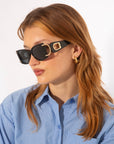 A person with long, wavy auburn hair is wearing For Art's Sake® Sculpture - Black acetate sunglasses, which feature 100% UVA & UVB protection and a gold D-logo on the temples. The person is dressed in a blue-striped button-up shirt and gold hoop earrings against a plain white background.