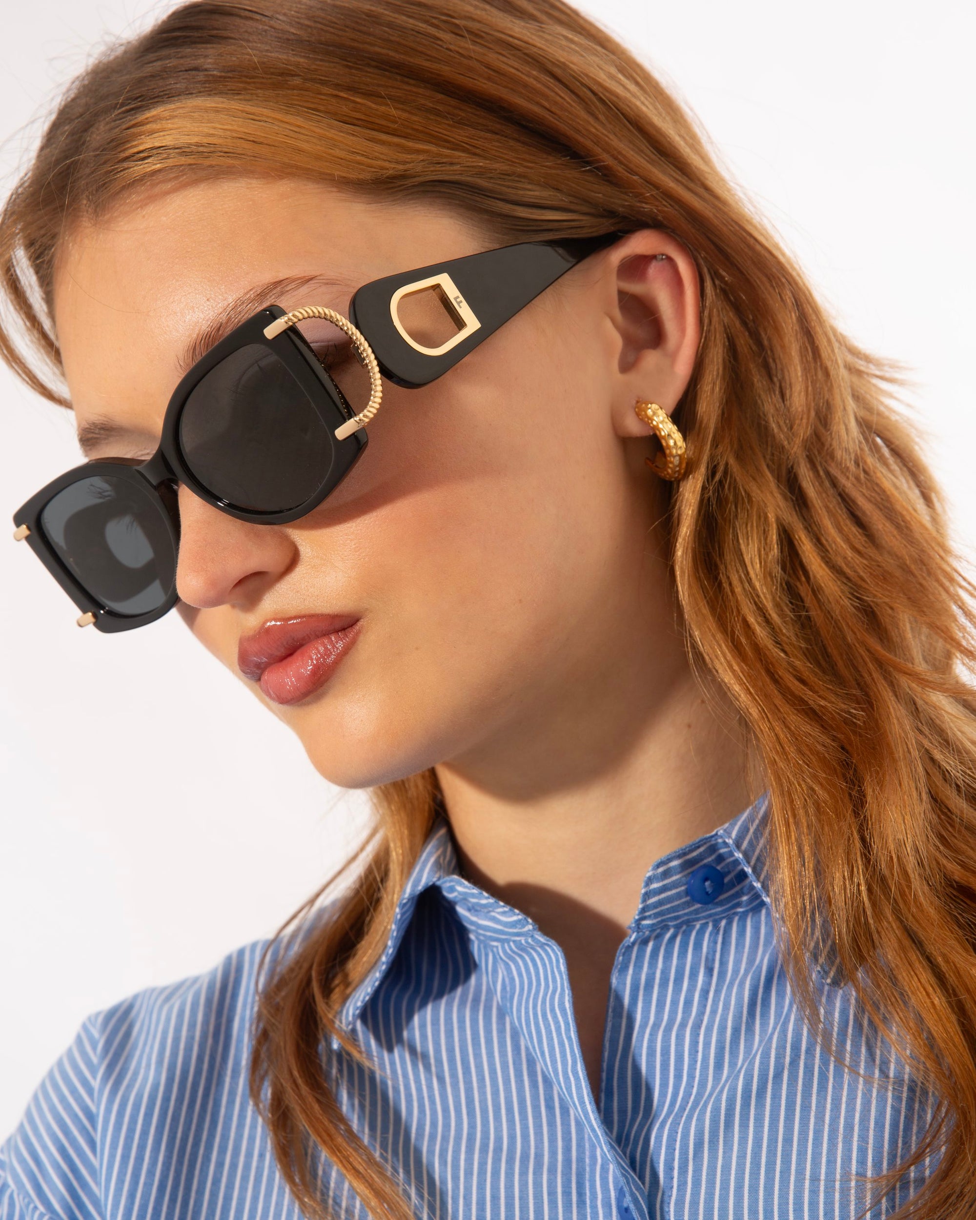 A person with long, wavy brown hair wears black, gold-plated For Art's Sake® Sculpture sunglasses with UV protection and a blue and white striped shirt. They also sport a gold hoop earring, and their head is tilted slightly to the left. The background is plain white.