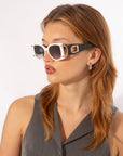 A person with light brown, shoulder-length hair is wearing For Art's Sake® Sculpture white-framed sunglasses with black temples and ultra-lightweight nylon lenses, a grey sleeveless top, and small dangling earrings. They are looking to the right against a plain white background.