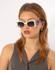 A woman with light brown, wavy hair is wearing a sleeveless gray top and Sculpture sunglasses by For Art's Sake® with white rims and dark lenses that provide 100% UVA & UVB protection. She has small, dangling earrings and is looking slightly off to the side. The background is plain white.