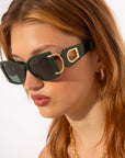 A young woman with long, light brown hair wearing oversized, dark green sunglasses with thick frames and gold accents. These chic For Art's Sake® Sculpture sunglasses provide 100% UVA & UVB protection. She also has gold hoop earrings and a gold necklace. The background is plain white, focusing attention on her stylish accessories.