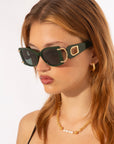 A woman with long, light brown hair is wearing dark green, rectangular sunglasses with gold accents on the temples. These For Art's Sake® Sculpture sunglasses offer 100% UVA & UVB protection and feature an Art Deco style. She also has gold hoop earrings and a gold chain necklace with pearl details, looking to the side against a plain white background.