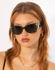 A person with auburn hair wears green Sculpture sunglasses by For Art's Sake® with gold accents, gold hoop earrings, and an 18-karat gold-plated necklace with small pearls. The background is plain white, emphasizing the person and their accessories.