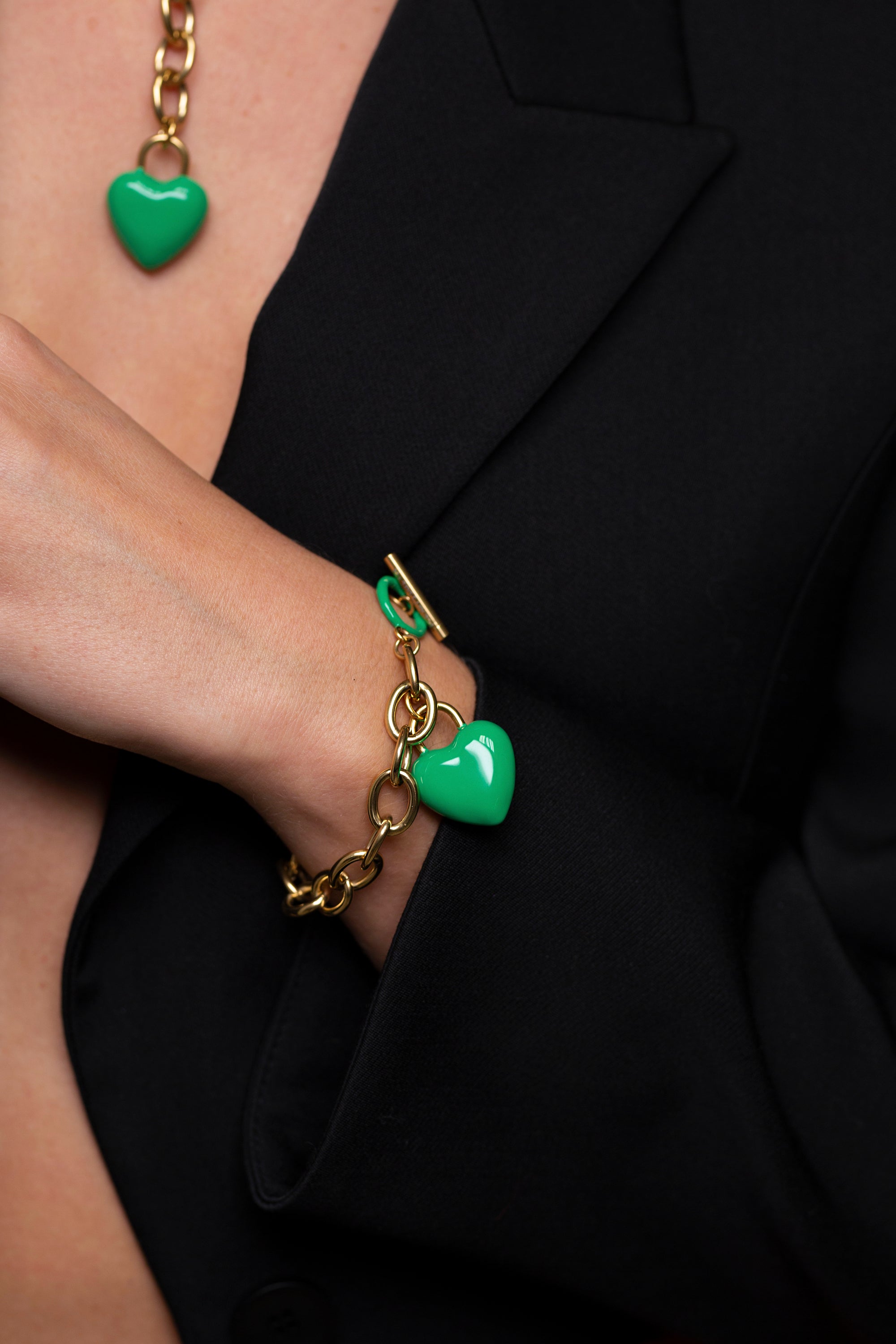 A person wearing a black blazer showcases The Kiss Bracelet by For Art's Sake®, an 18k gold chain bracelet adorned with an enamel-covered heart charm. A matching necklace with a green heart pendant is partially visible. The person's hand is tucked into the blazer.