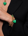 A person wearing a black blazer showcases The Kiss Bracelet by For Art's Sake®, an 18k gold chain bracelet adorned with an enamel-covered heart charm. A matching necklace with a green heart pendant is partially visible. The person's hand is tucked into the blazer.