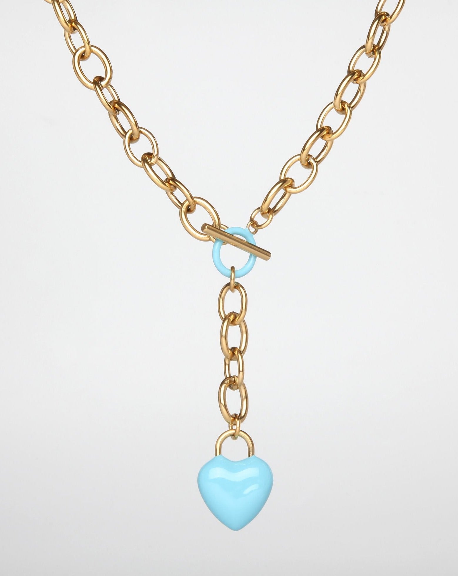 A gold necklace featuring interlocking links has a toggle clasp and a dangling light blue, enamel-coated heart-shaped pendant. The heart is shiny and smooth, contrasting with the metallic texture of the chain. Reminiscent of "The Kiss," it evokes timeless romance against a plain white background. This is the For Art's Sake® The Kiss Necklace Pink.
