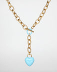 A gold necklace featuring interlocking links has a toggle clasp and a dangling light blue, enamel-coated heart-shaped pendant. The heart is shiny and smooth, contrasting with the metallic texture of the chain. Reminiscent of "The Kiss," it evokes timeless romance against a plain white background. This is the For Art's Sake® The Kiss Necklace Pink.