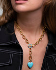 Close-up of a person wearing For Art's Sake® The Kiss Necklace Pink with a large enamel-coated heart pendant and a toggle clasp. The person is dressed in a black garment with white stitching. The necklace stands out against the dark background of the clothing.