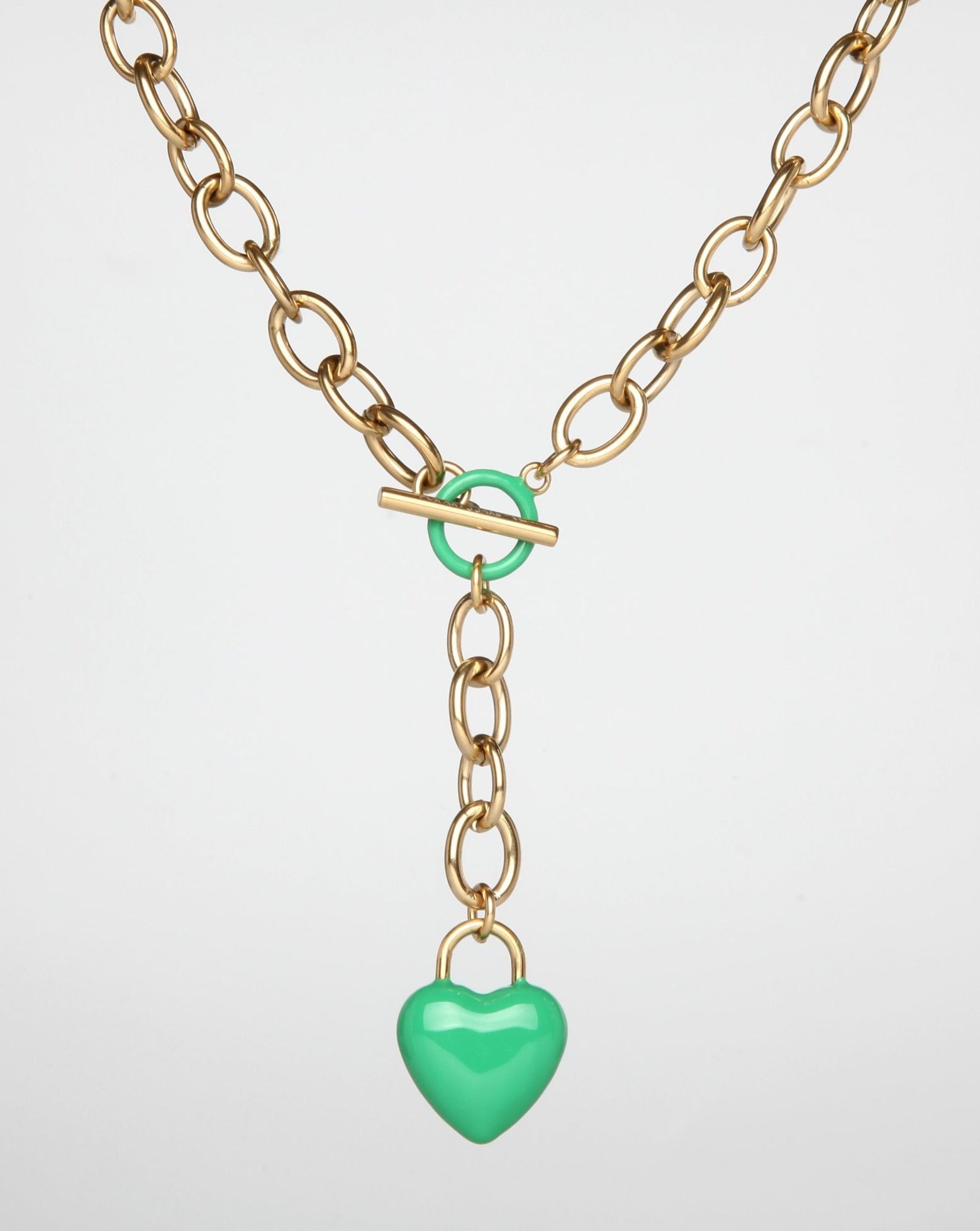 A gold necklace with a clasp and a green enamel-coated heart-shaped pendant. The Kiss Necklace Pink by For Art's Sake® features large interlocking links and a toggle closure, with the vibrant heart hanging at the center of the chain on a short length of additional links.